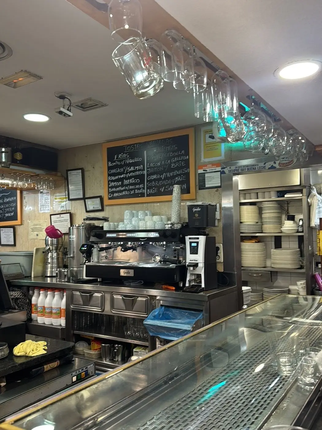 Barra de la cafetería con la cafetera, la pizarra de tostas y copas colgadas del techo