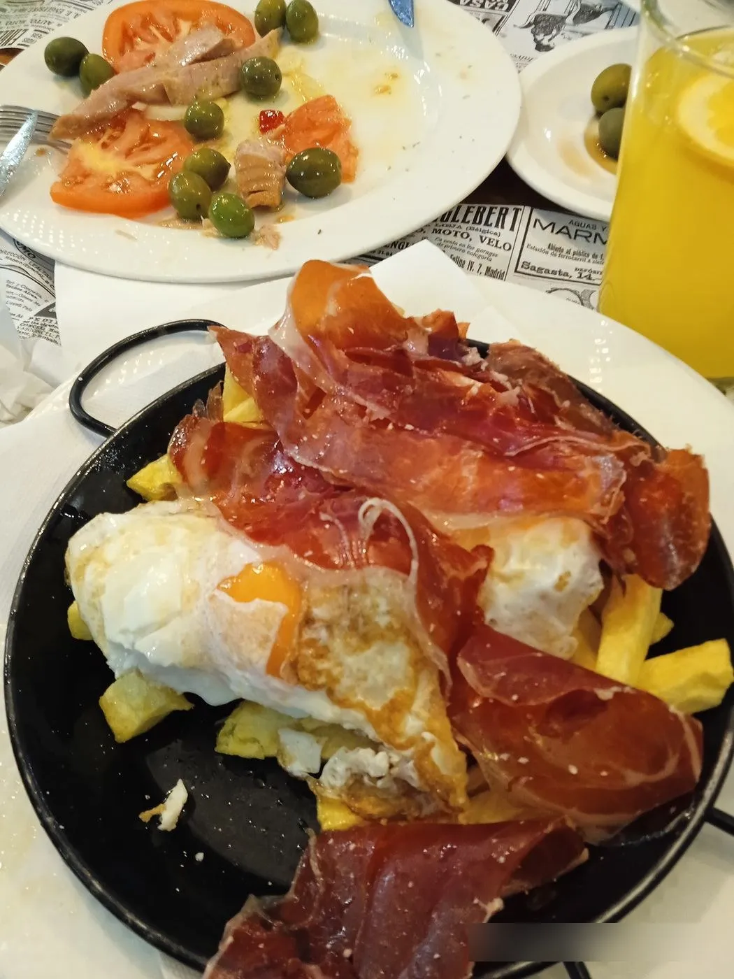 Huevos fritos con patatas y jamón en una sartén negra, junto a un zumo de naranja